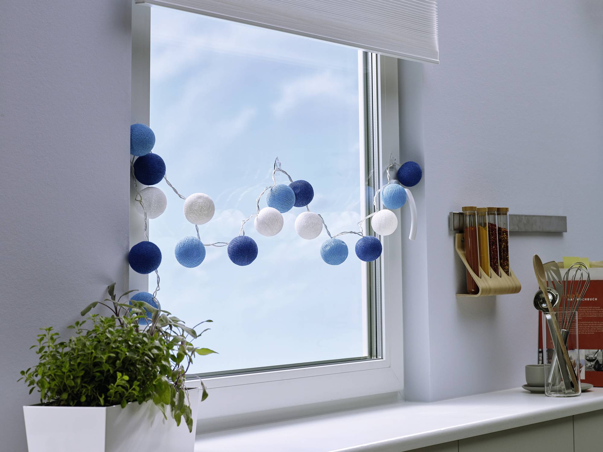 Fairy lights made of blue and white baubles hang at a window. Beside them are kitchen utensils and a plant against a blue sky.