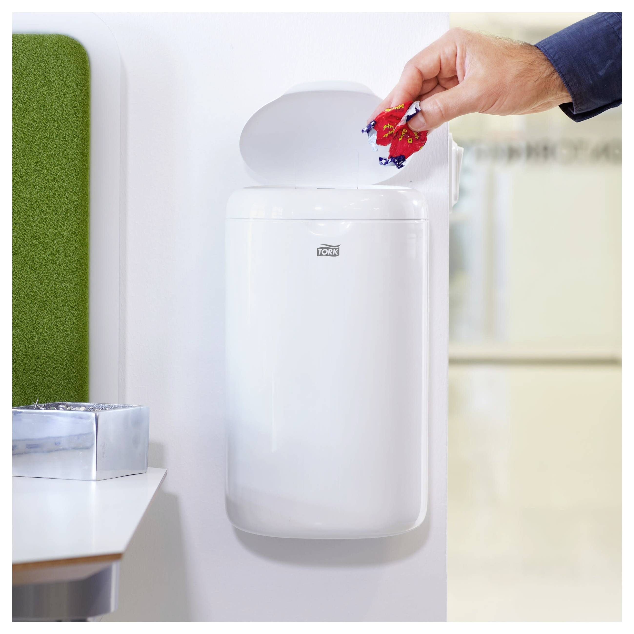 Hand drops crumpled paper into a white wall-mounted Tork waste bin in a bright restroom.