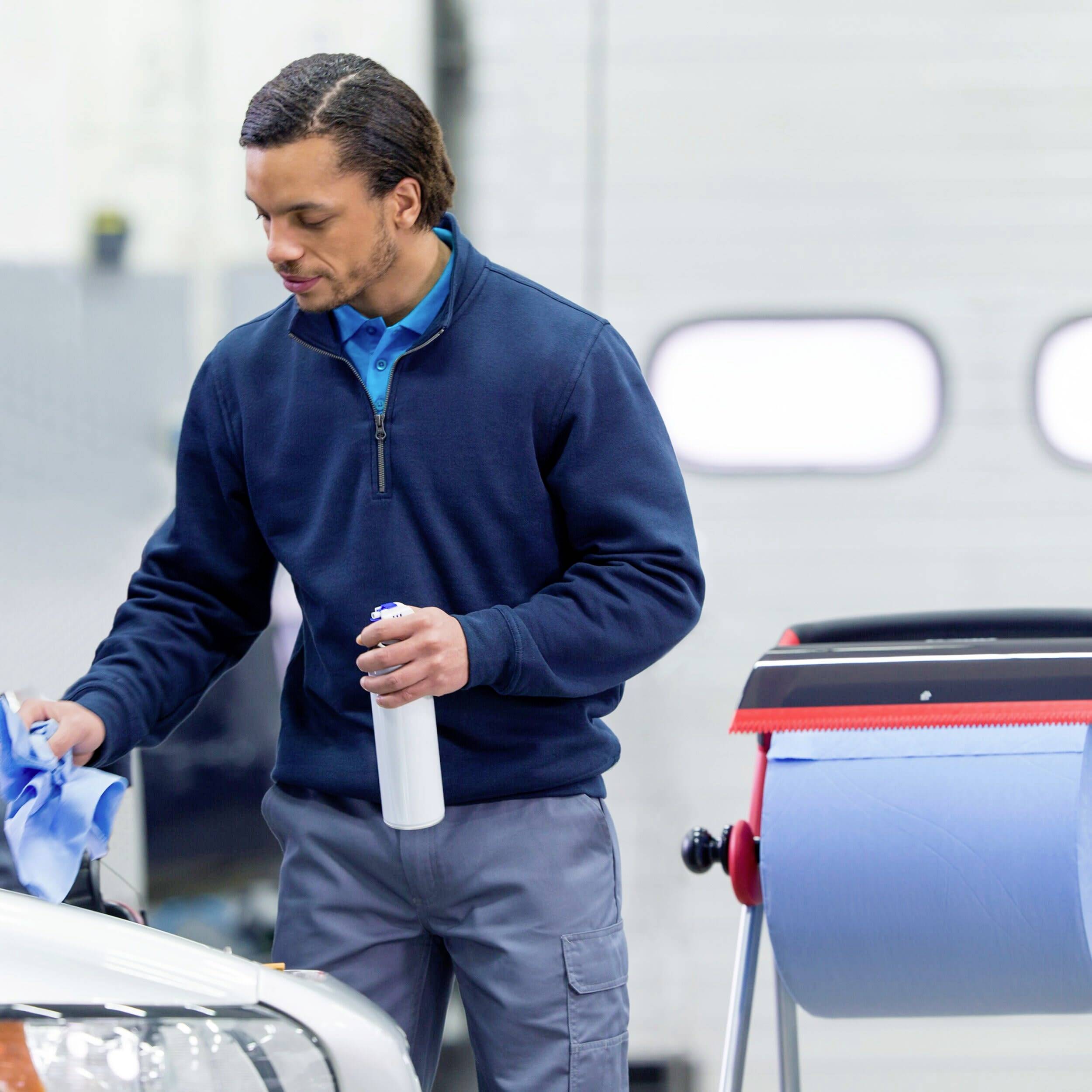 A man is cleaning a headlight cover with a cloth and spray in a workshop. Car doors can be seen in the background.