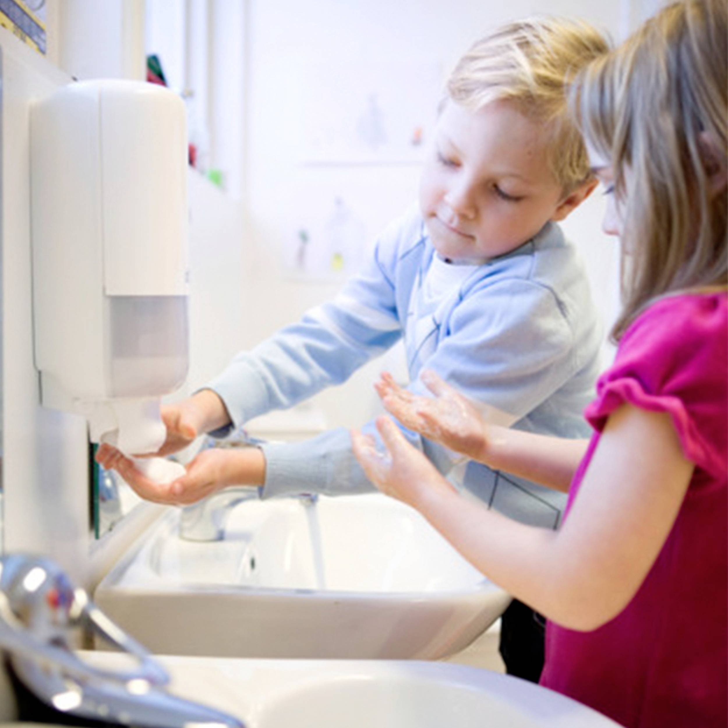 A boy and a girl are washing their hands at a sink, using soap from a dispenser.