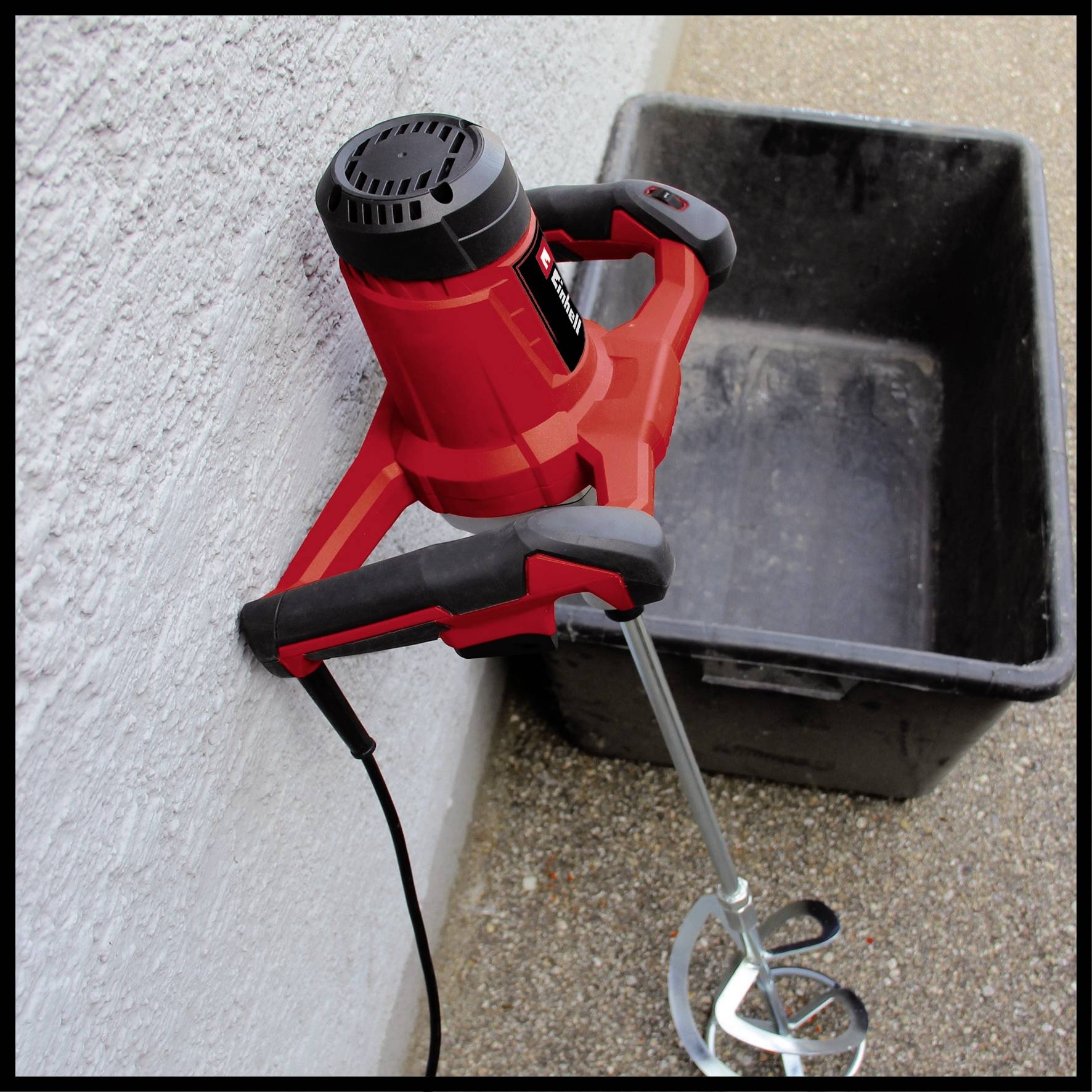 A red electric whisk is leaning against a wall next to an empty, black rectangular container on a gravel floor.