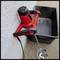 'A red electric concrete mixer with a stirring rod is leaning against a wall next to a black plastic trough on a concrete floor.'