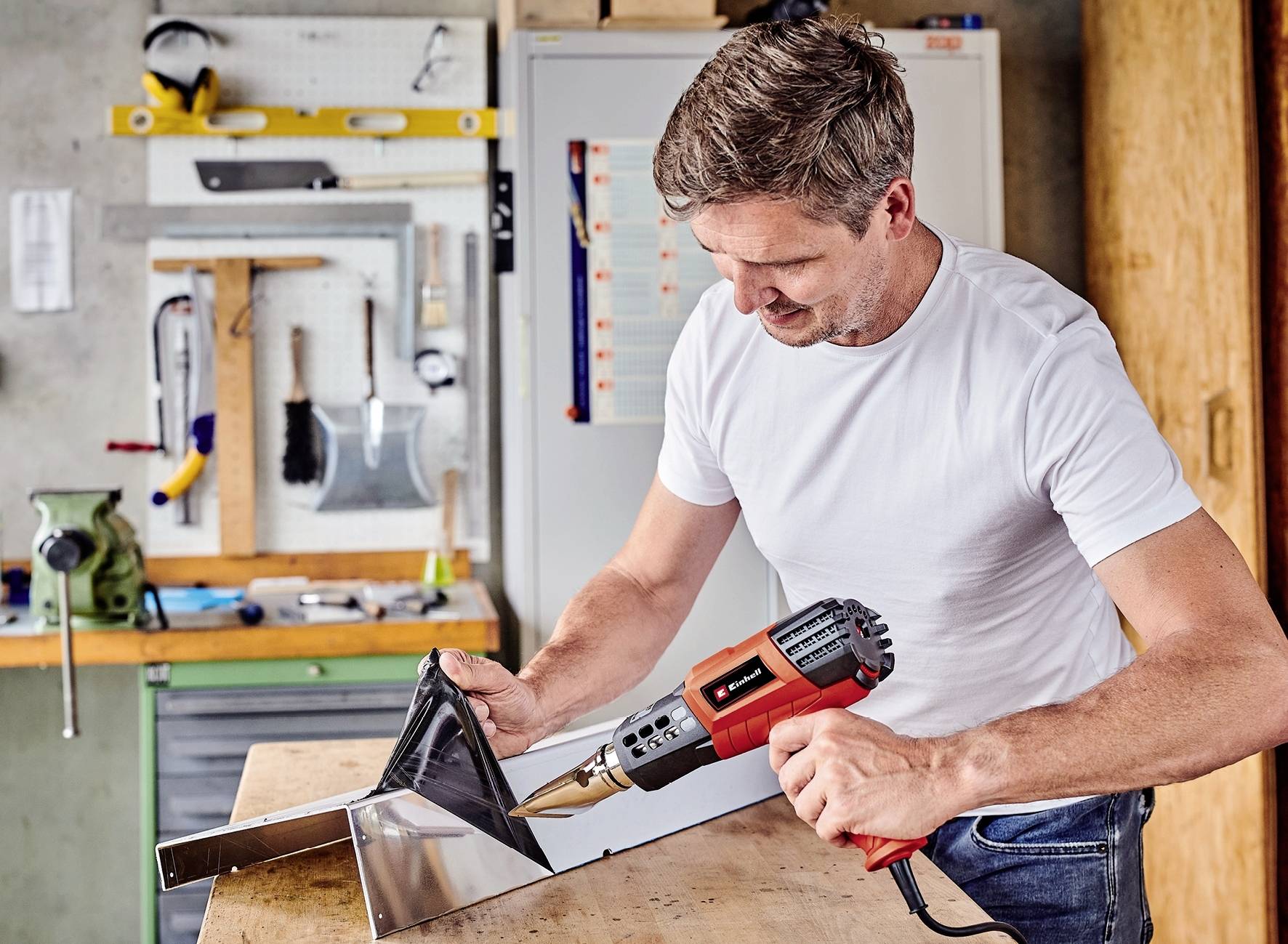 A man in a workshop is working on a metal piece using a heat gun. Tools are visible in the background.