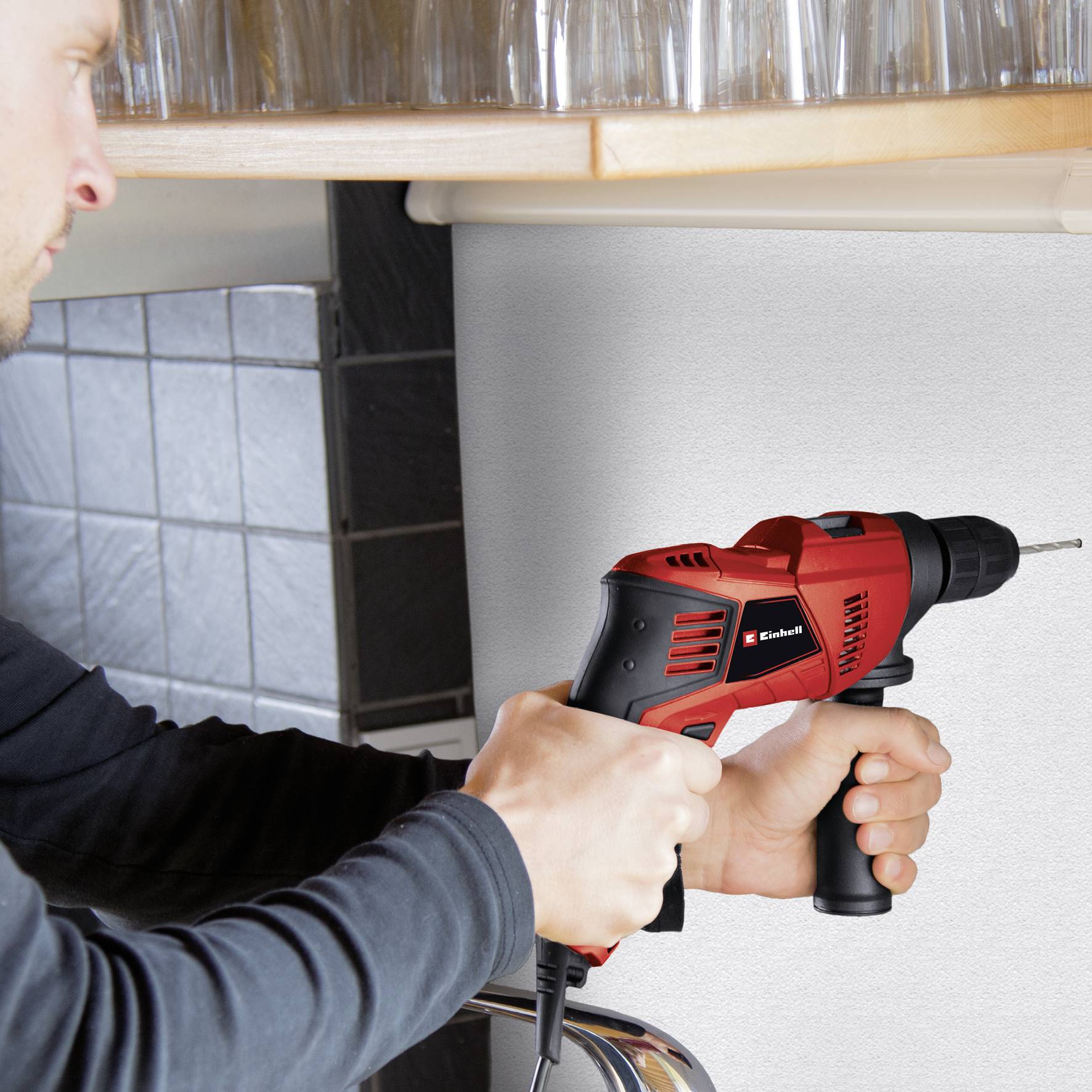 A person is drilling into a white wall with a red drill to create a hole for a fixing. Kitchen utensils are visible in the background.
