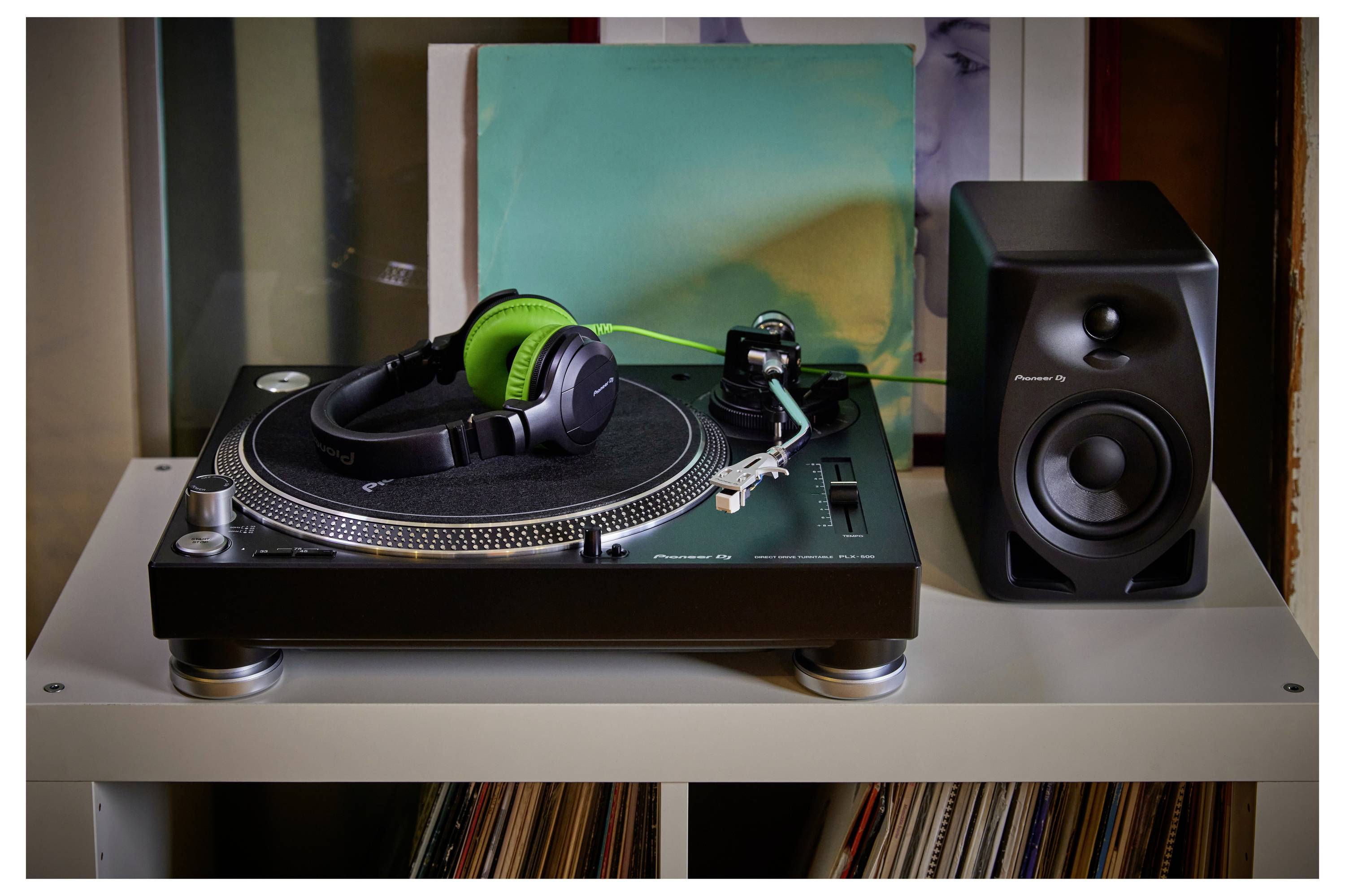 A turntable with green-and-black headphones resting on it beside a black speaker, with records stored below on the shelf.
