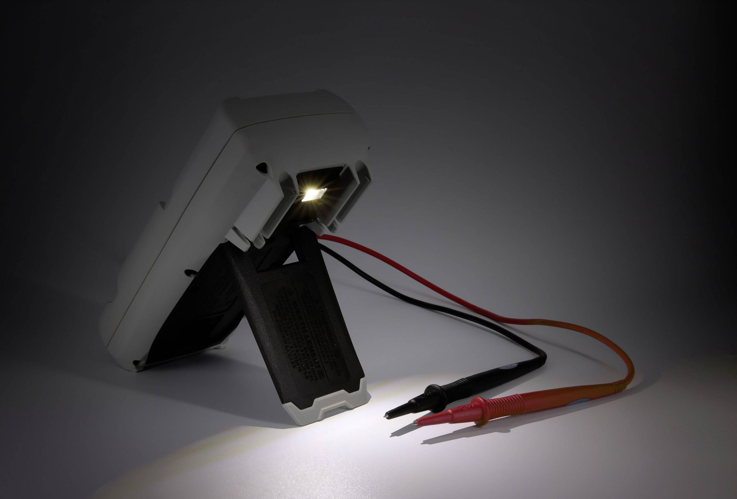 A multimeter with an illuminated display is resting on a surface. Two test leads in red and black are protruding.