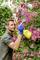 A person is spraying water onto a flowering rose bush in the garden. They are wearing blue gardening gloves and holding a yellow spray bottle, with a focused expression.