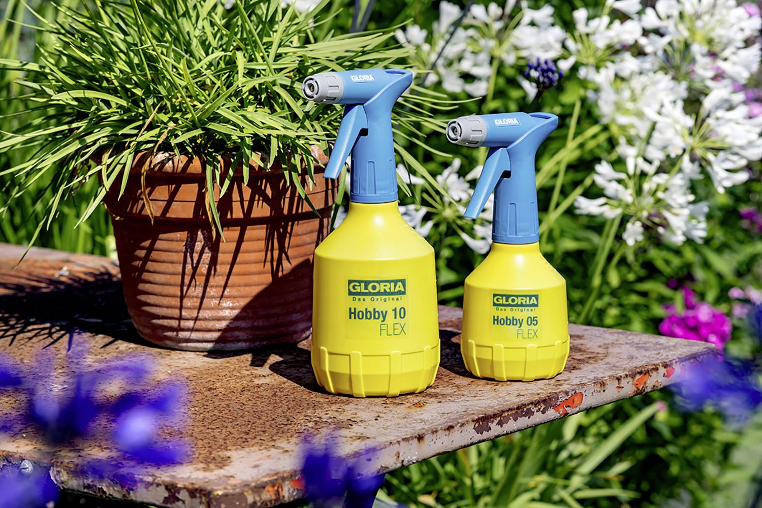 Two yellow spray bottles from the Gloria brand, 'Hobby 10 FLEX' and 'Hobby 05 FLEX', are standing on an old table next to a flower pot in the garden.