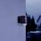 A black outdoor motion sensor camera on the wall of a building at dusk. Background shows blurred garden area.
