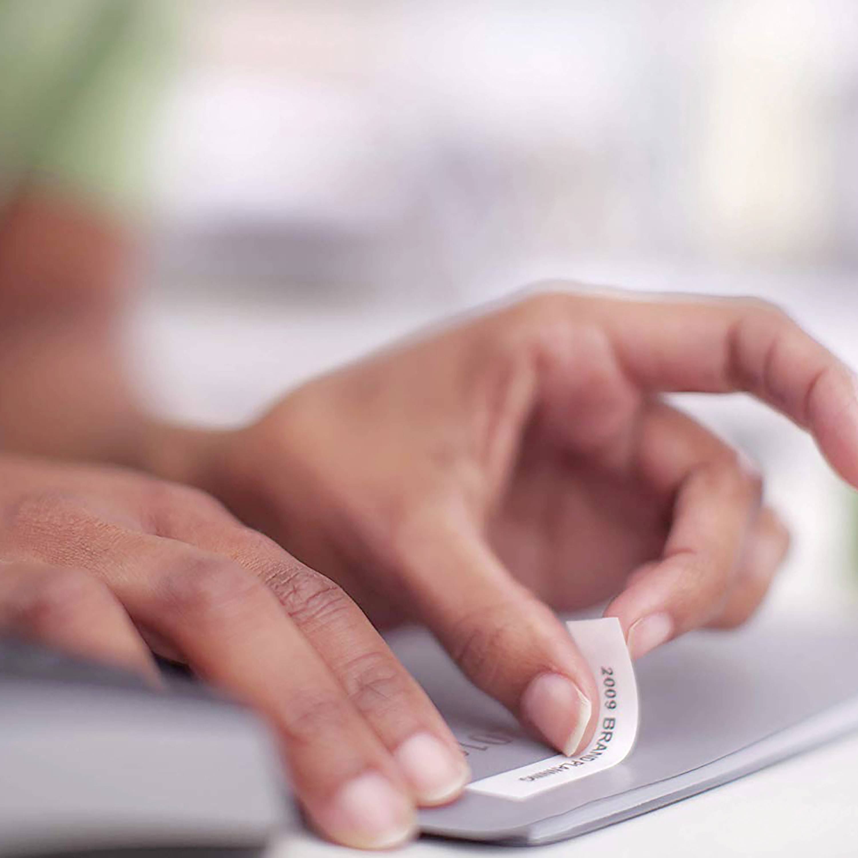 A hand is pressing a button on a white label printer. Blurred objects are visible in the background.