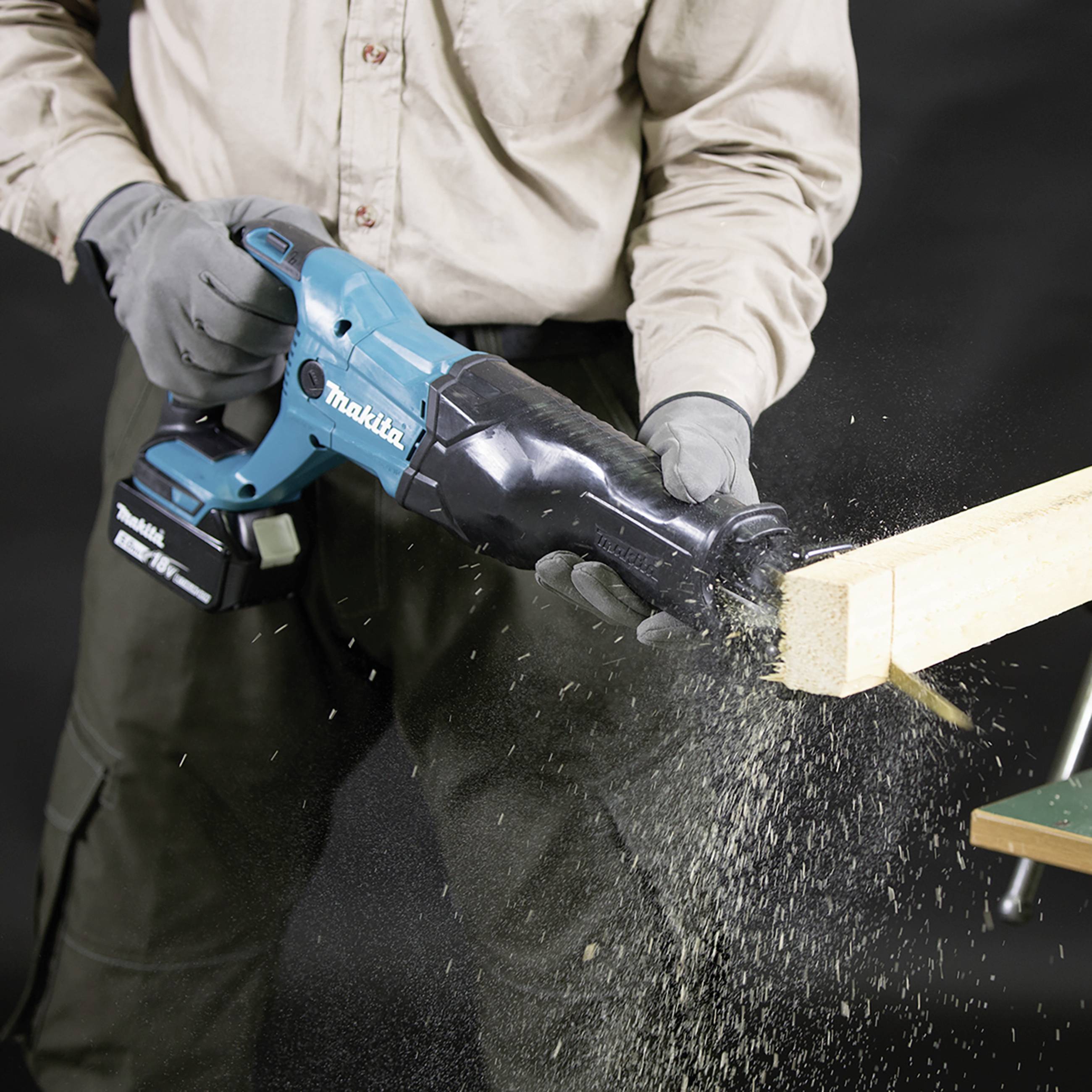 A person is cutting a piece of wood with a blue electric reciprocating saw. Wood shavings are flying. The saw is a Makita brand.