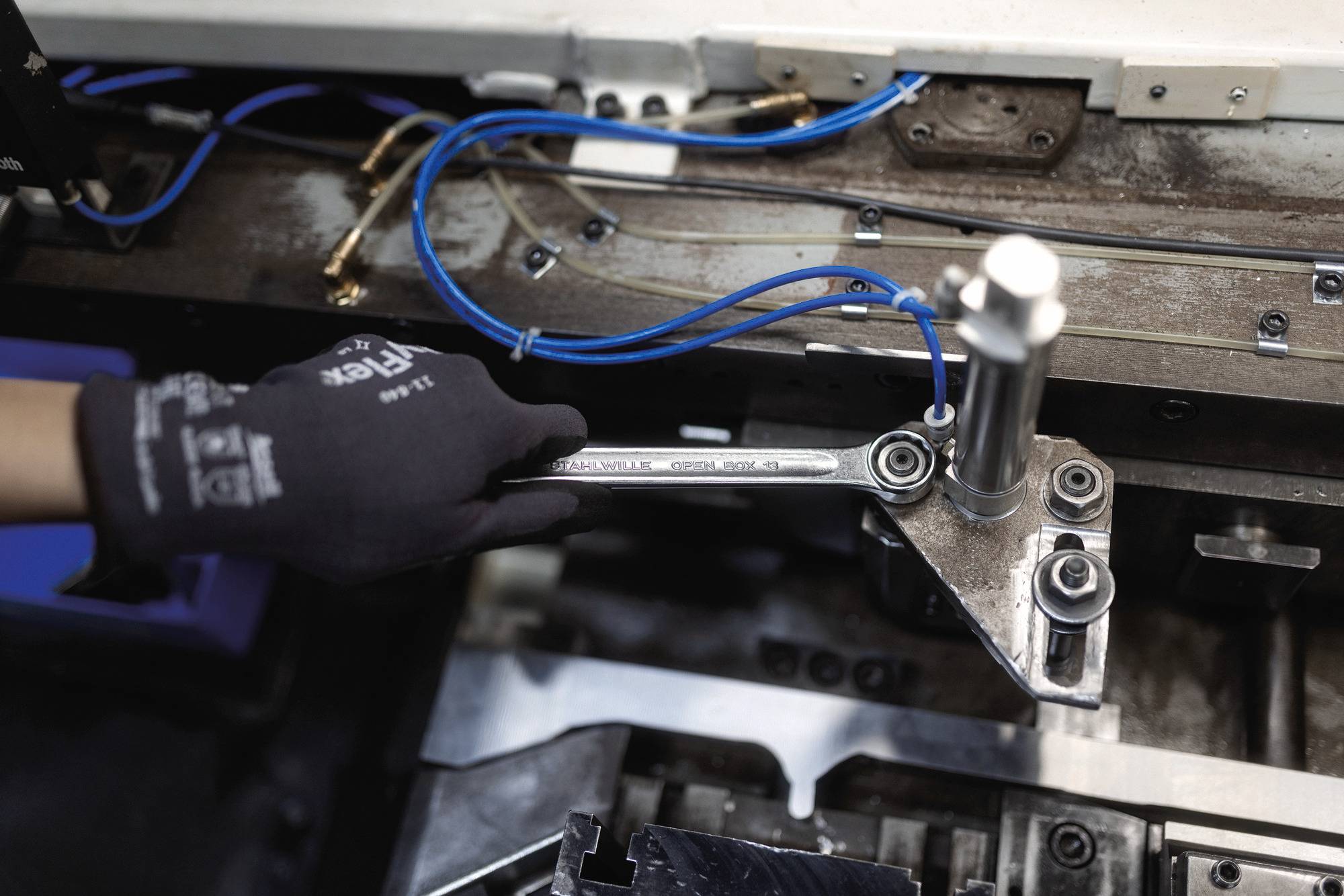 A gloved hand adjusts a mechanical component with a wrench on an industrial machine, connected by blue wires, in a factory setting.