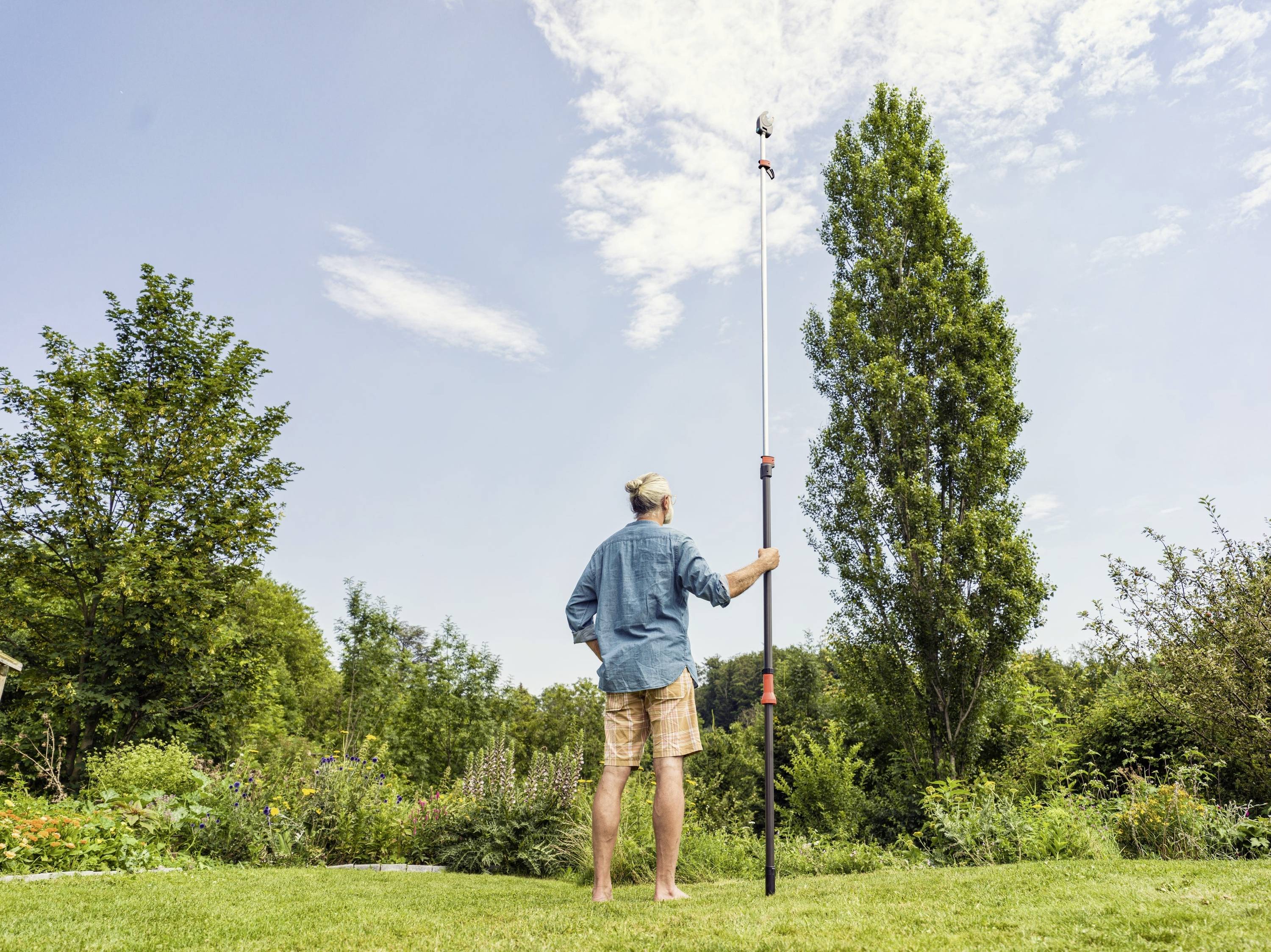 A man is standing in the garden, holding a long pole with a camera attached to capture photographs from above.