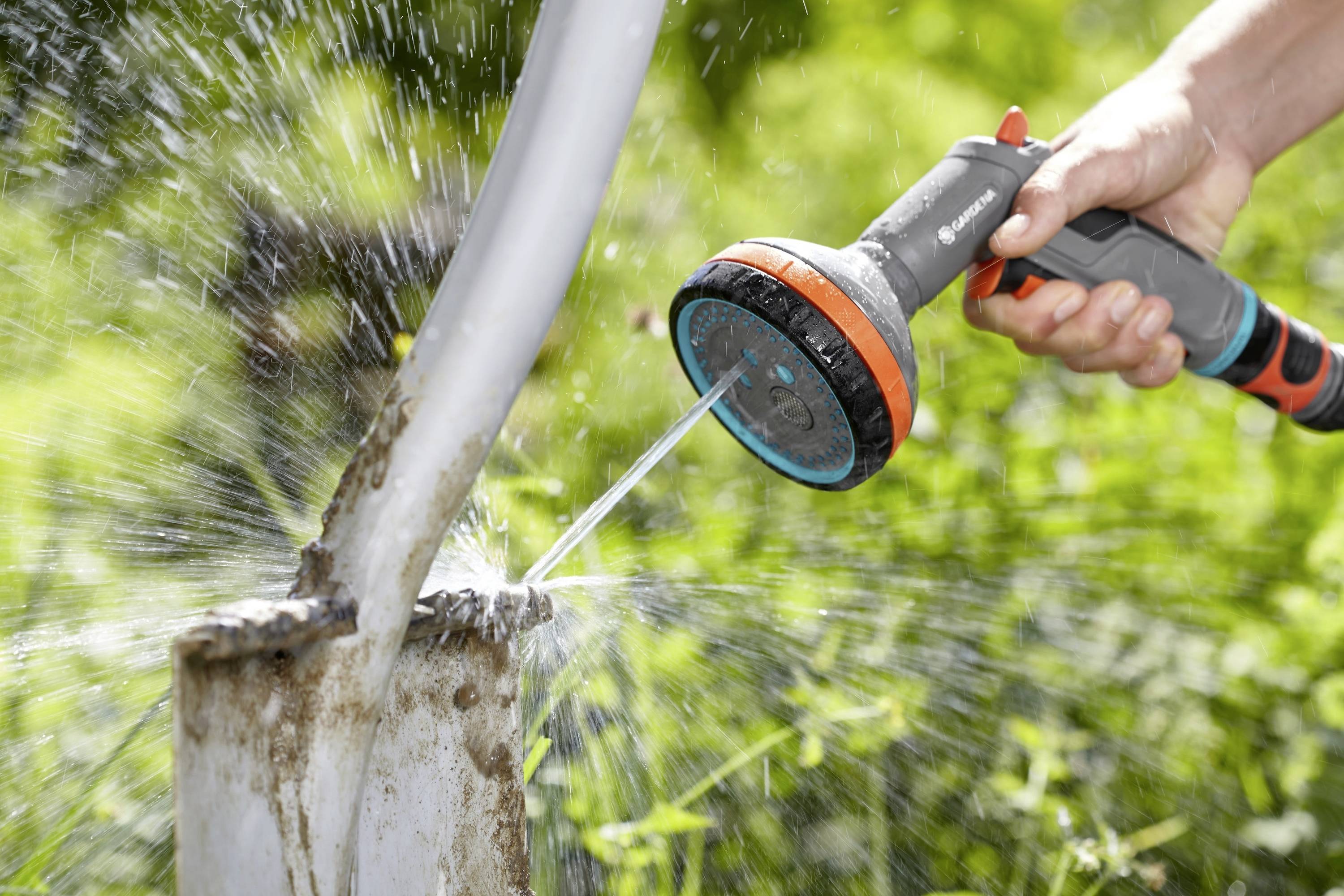 A person is holding a garden hose with a spray nozzle and washing a muddy metal pipe. Water is powerfully spraying forth.