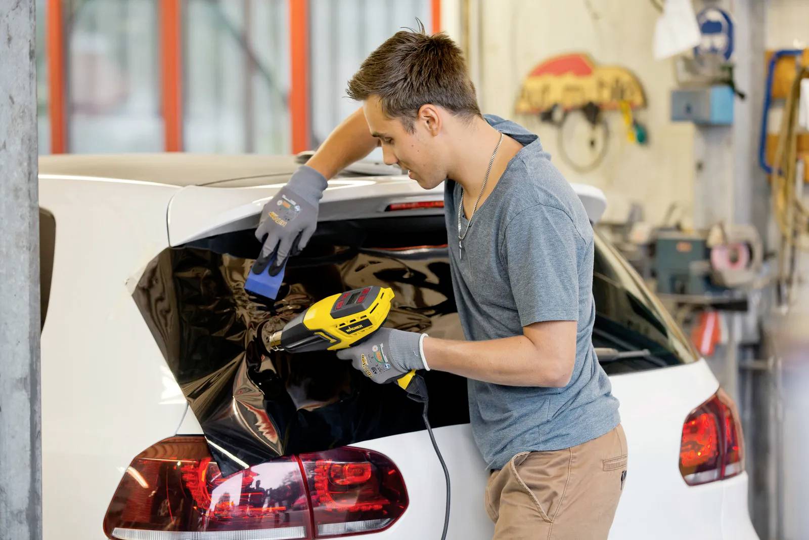 A man in a workshop environment is applying tinted film to the rear window of a white car.