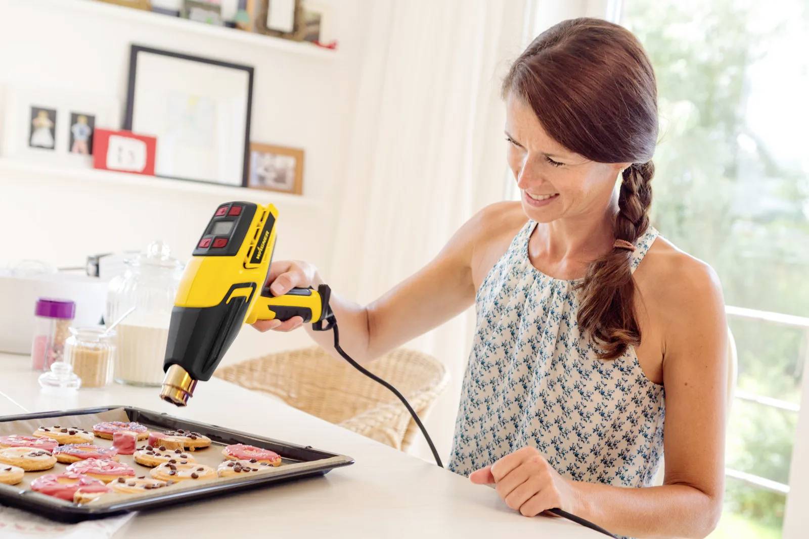 A woman is decorating colourful biscuits on a baking tray with a piping device in a bright kitchen.