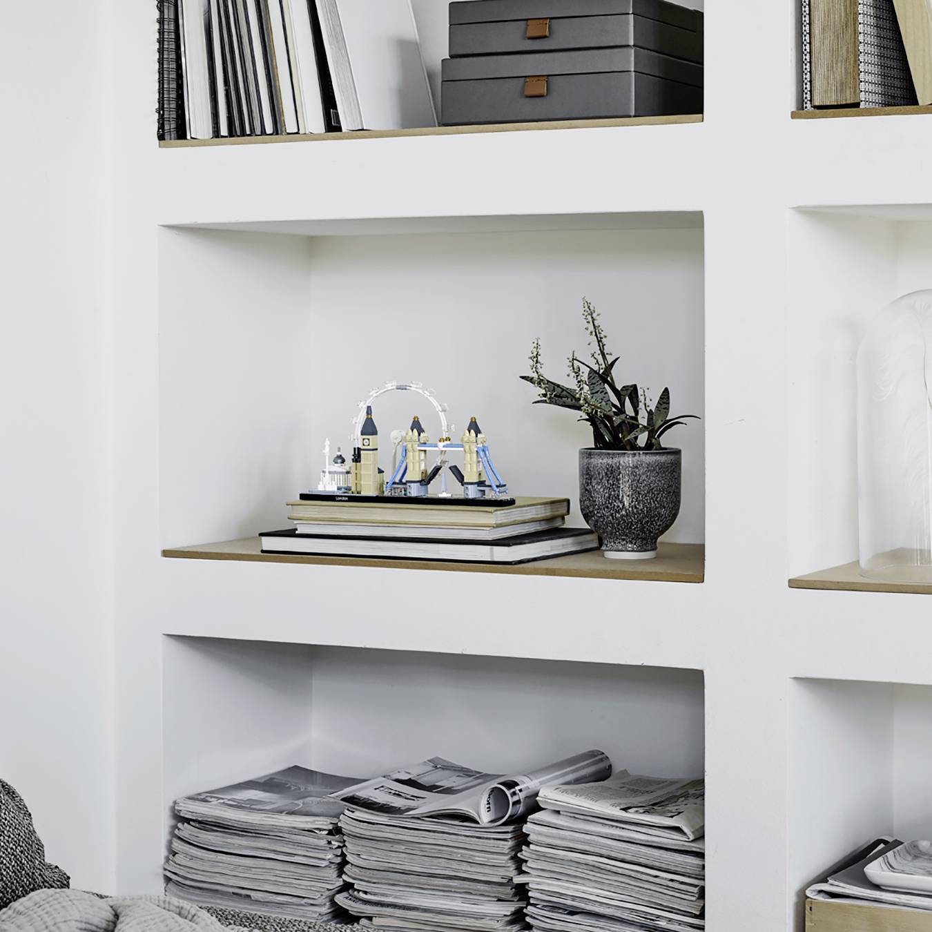 A white shelving unit with decorative boxes, a model of the London Eye, plants, and stacked magazines.