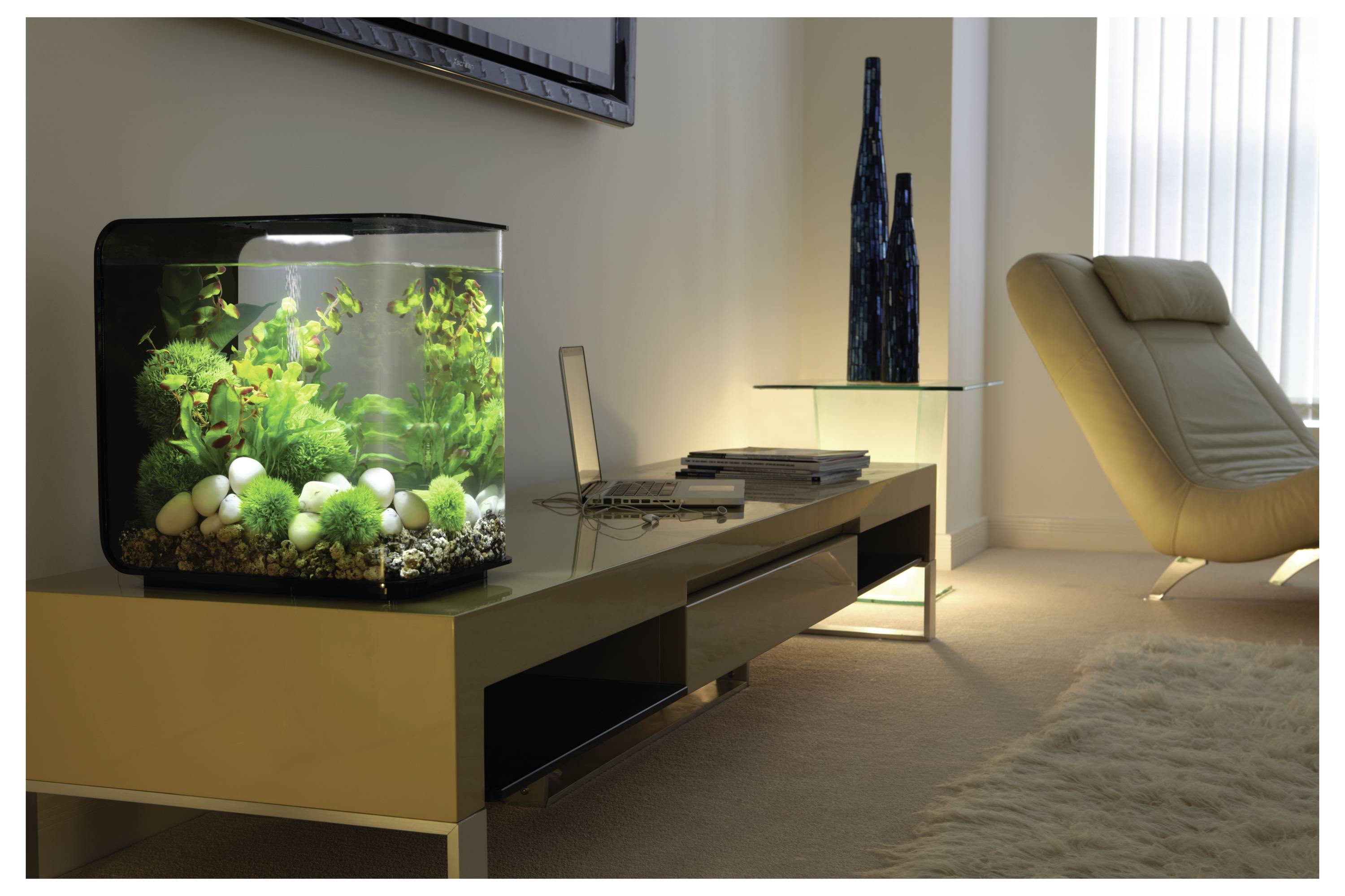 A modern living room features a sleek beige lounger, a low-profile table with a laptop, and a lit fish tank with vibrant green plants.