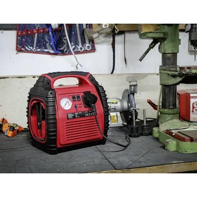 A red air compressor sits on a workbench in a workshop. Tools and a drill are visible in the background.