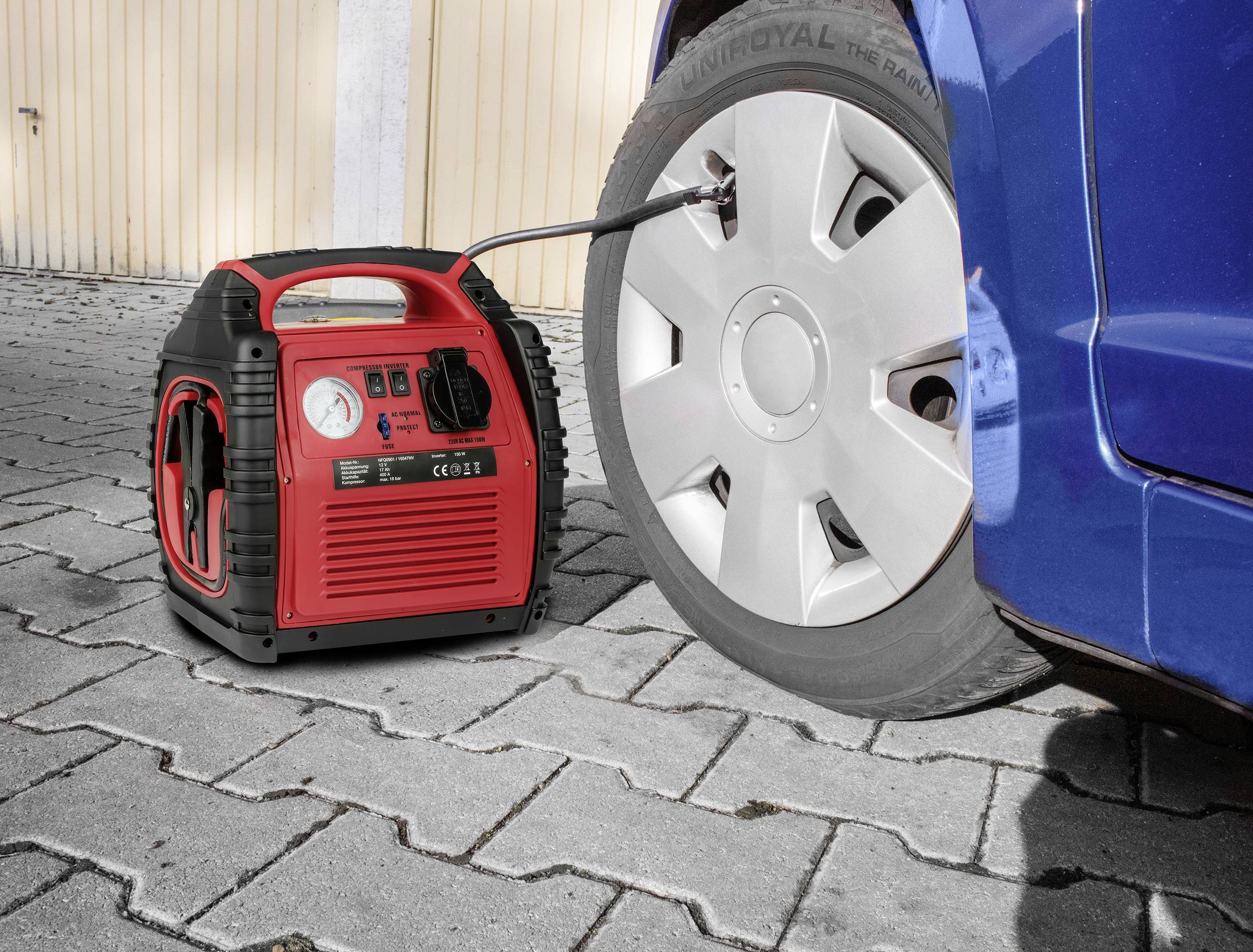 A red compressor is pumping up a car tyre. The vehicle is standing on paved ground next to a wooden wall.