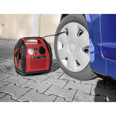 A red compressor is pumping up a car tyre. The vehicle is standing on paved ground next to a wooden wall.