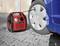 A red compressor is pumping up a car tyre. The vehicle is standing on paved ground next to a wooden wall.