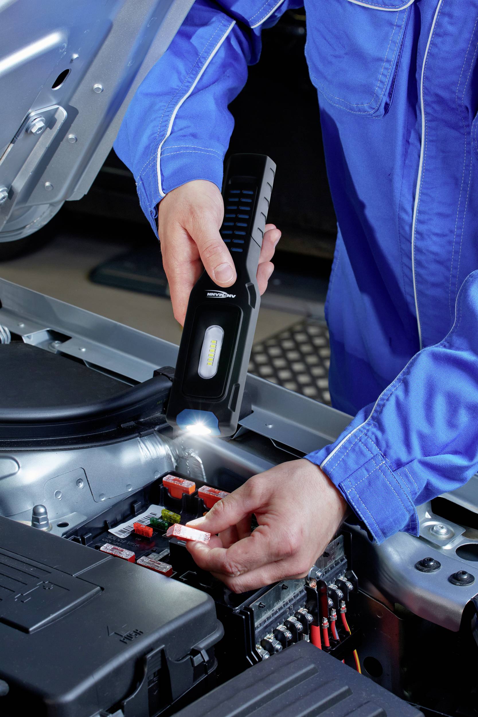 A person in blue workwear is checking the fuses in an open car engine using a torch.