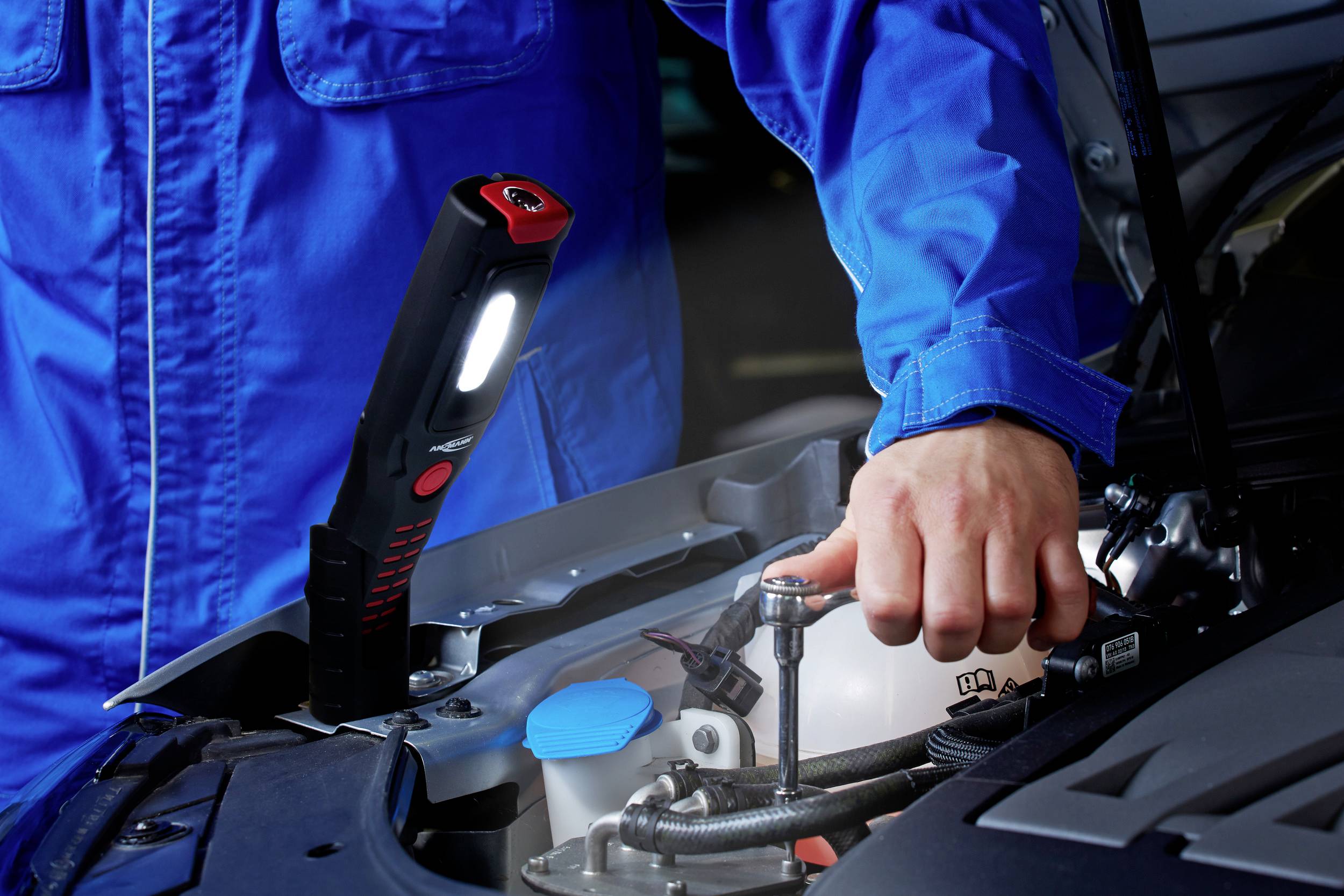 A person in blue workwear is repairing a car with a spanner. A workshop lamp illuminates the engine compartment.