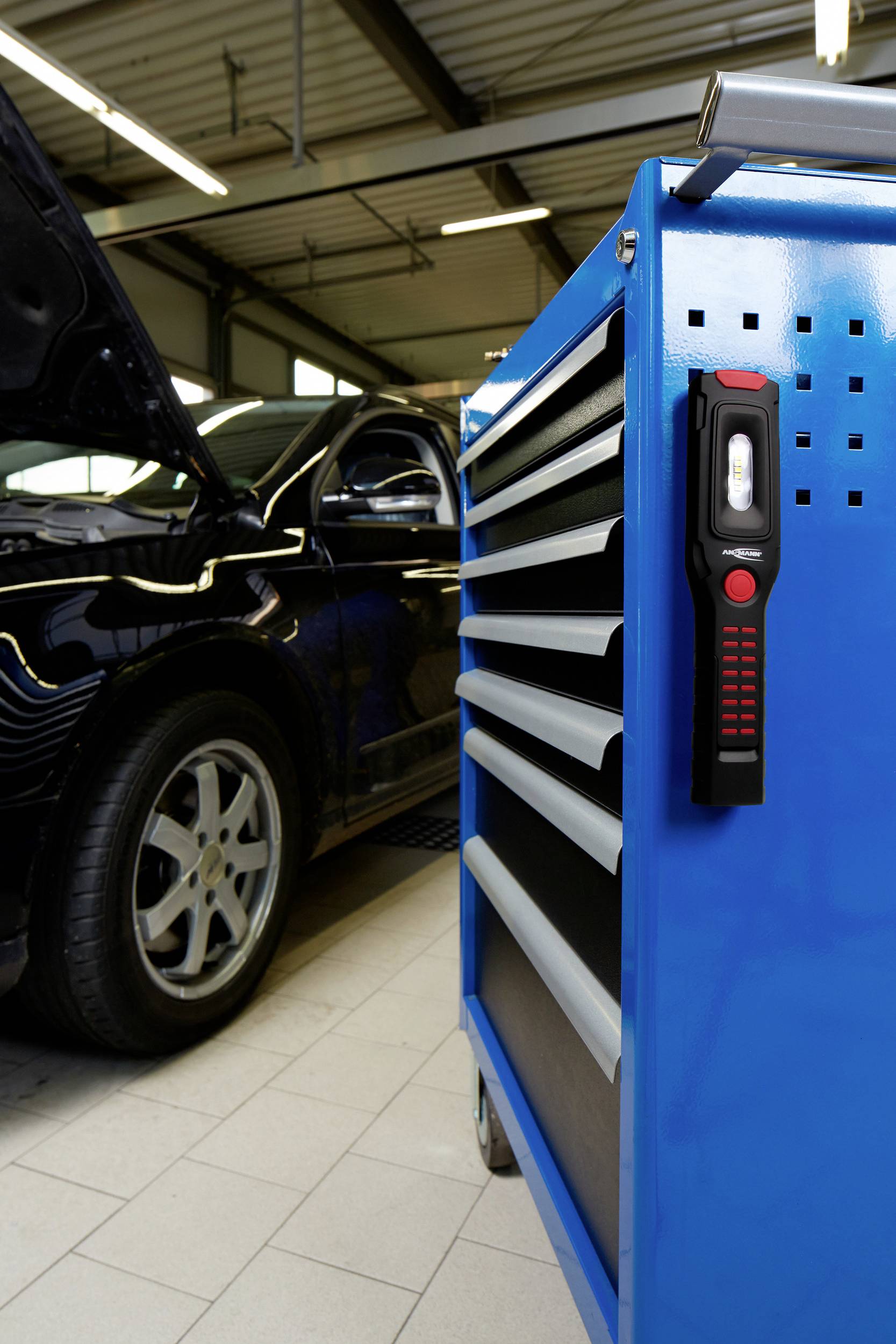 A black car with its bonnet open is parked in a garage. In the foreground, a blue tool trolley with a lamp is visible.