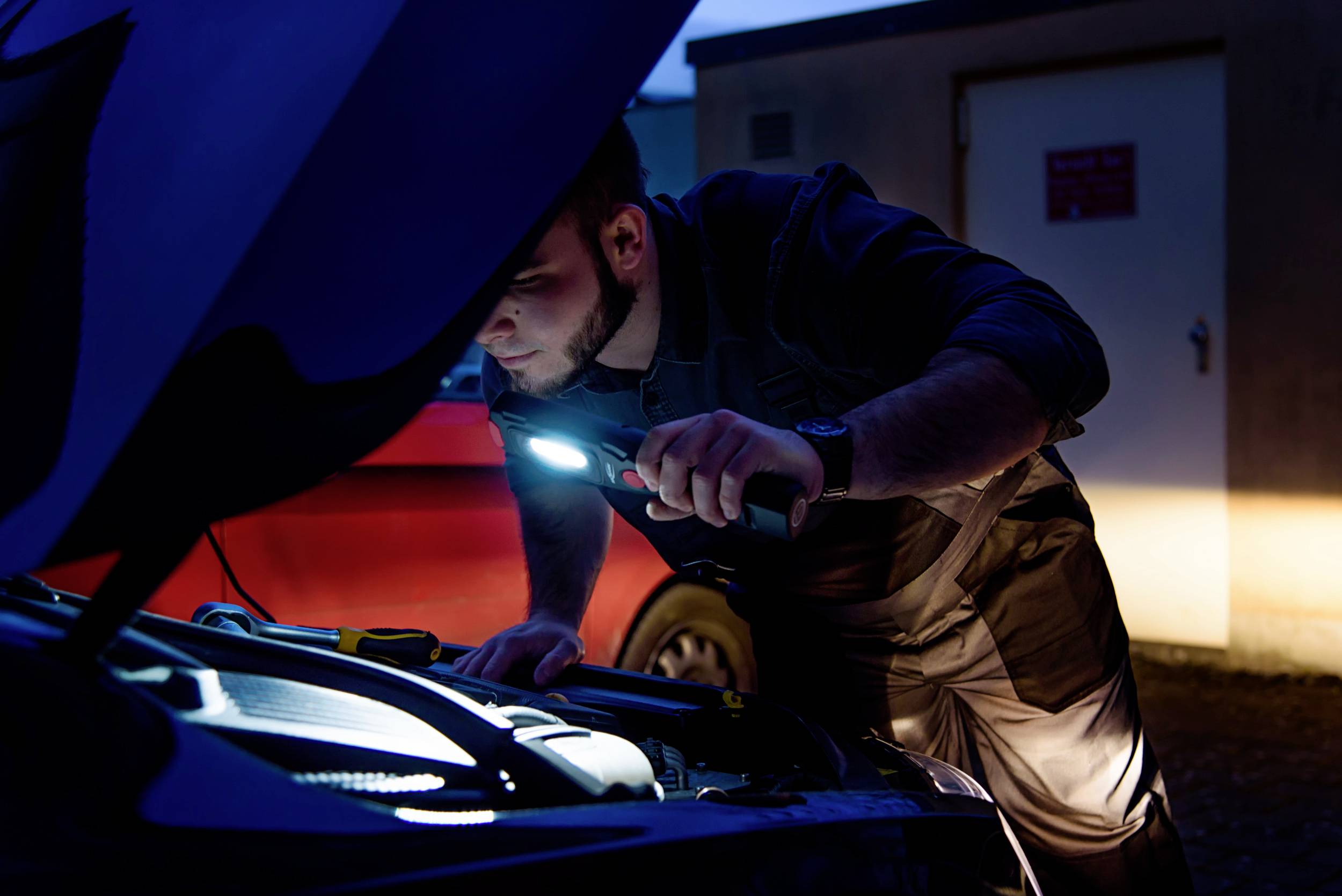 A person is examining the engine compartment of a car with a torch at dusk.