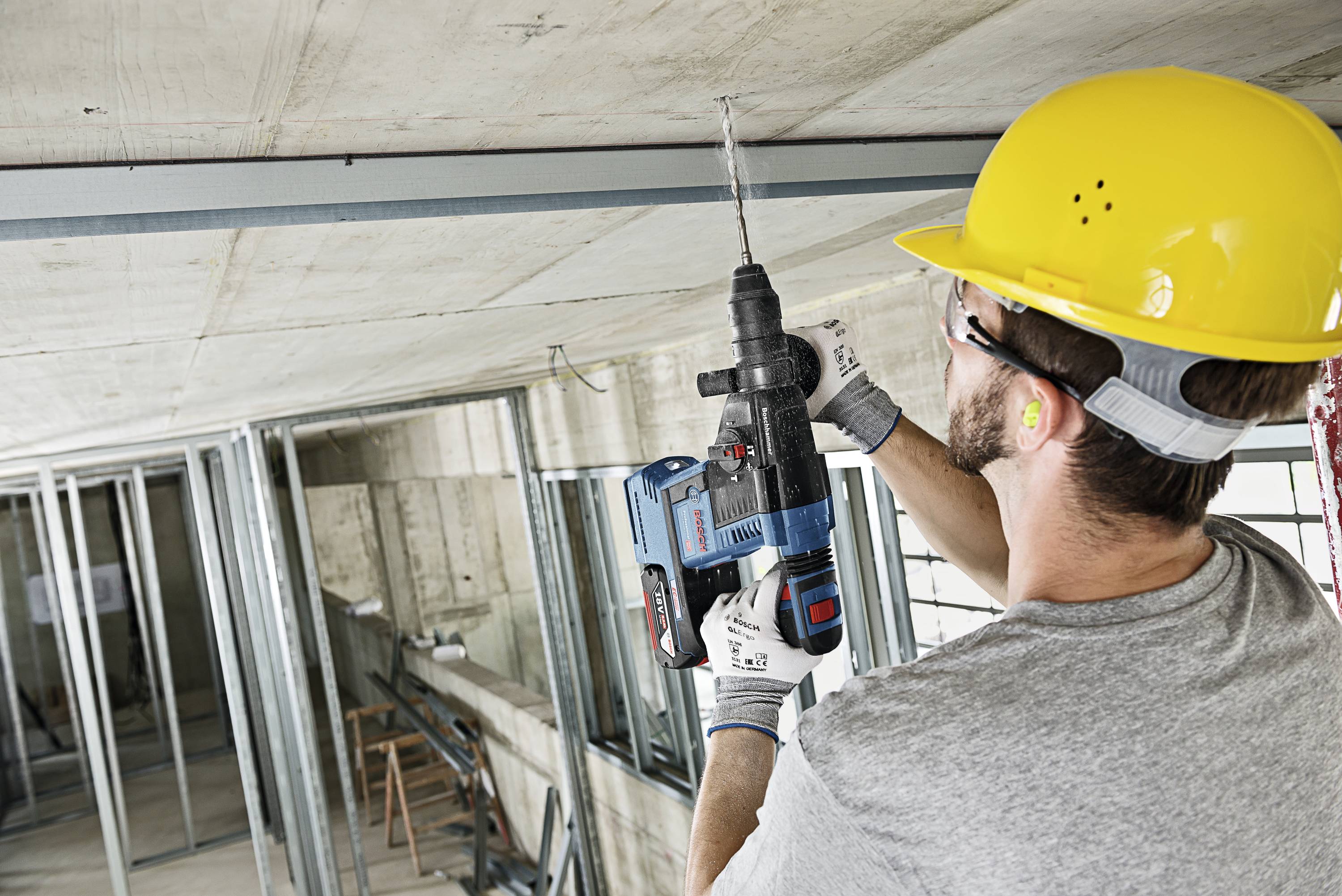 A worker is drilling into the ceiling of a construction site using an electric drill. He is wearing a yellow hard hat and ear plugs.