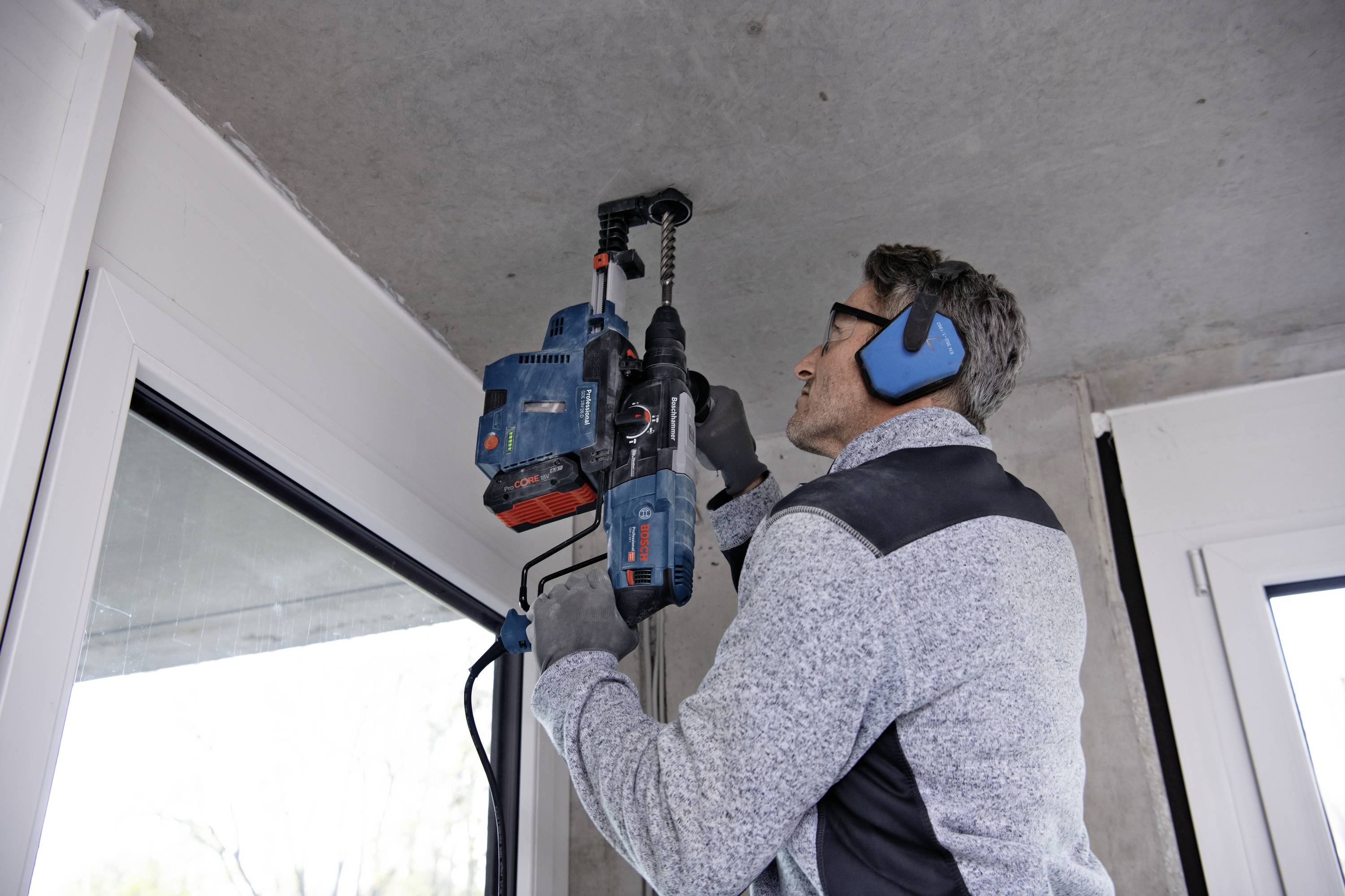 A man is using a drill to bore into the concrete ceiling above a window. He is wearing ear protection and gloves.