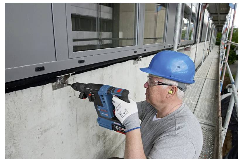 A worker in a blue hard hat and gloves uses a power drill on an exterior wall beneath windows. He wears safety glasses and ear protection.