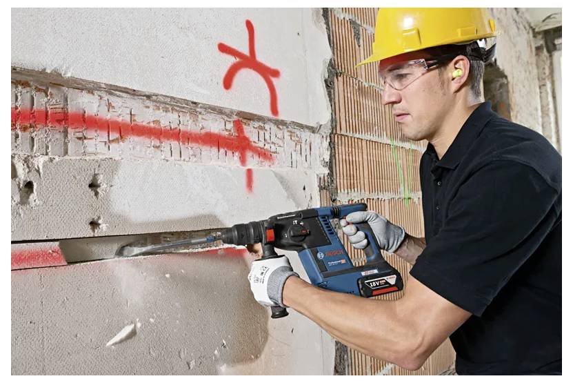 A construction worker wearing protective gear uses a power tool to cut through a concrete wall marked with red spray paint lines.