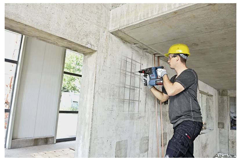 A construction worker in a yellow hard hat uses a power drill on a concrete wall. The building's unfinished interior is visible.