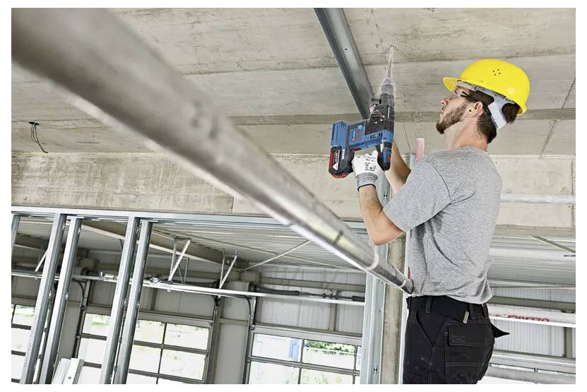 A worker in a hard hat uses a power drill on a construction site ceiling, showing active construction work indoors.