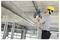 A worker in a hard hat uses a power drill on a construction site ceiling, showing active construction work indoors.