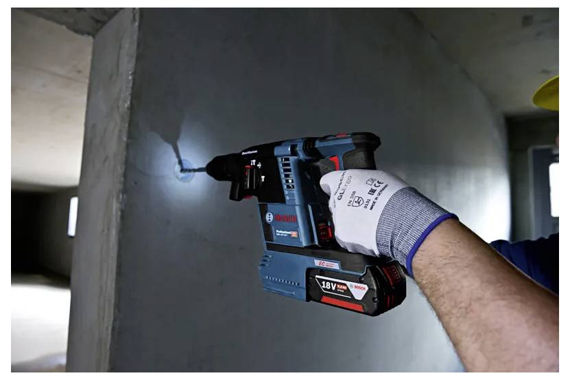 A person wearing gloves uses a blue and red power drill to make a hole in a concrete wall inside a building under construction.