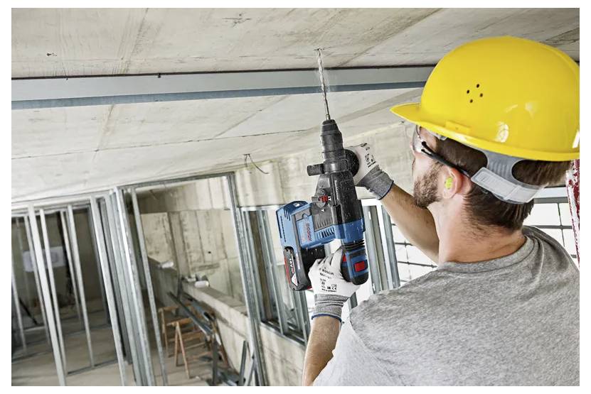 Worker wearing a yellow hard hat uses a power drill on a concrete ceiling in a construction site, surrounded by metal framing.
