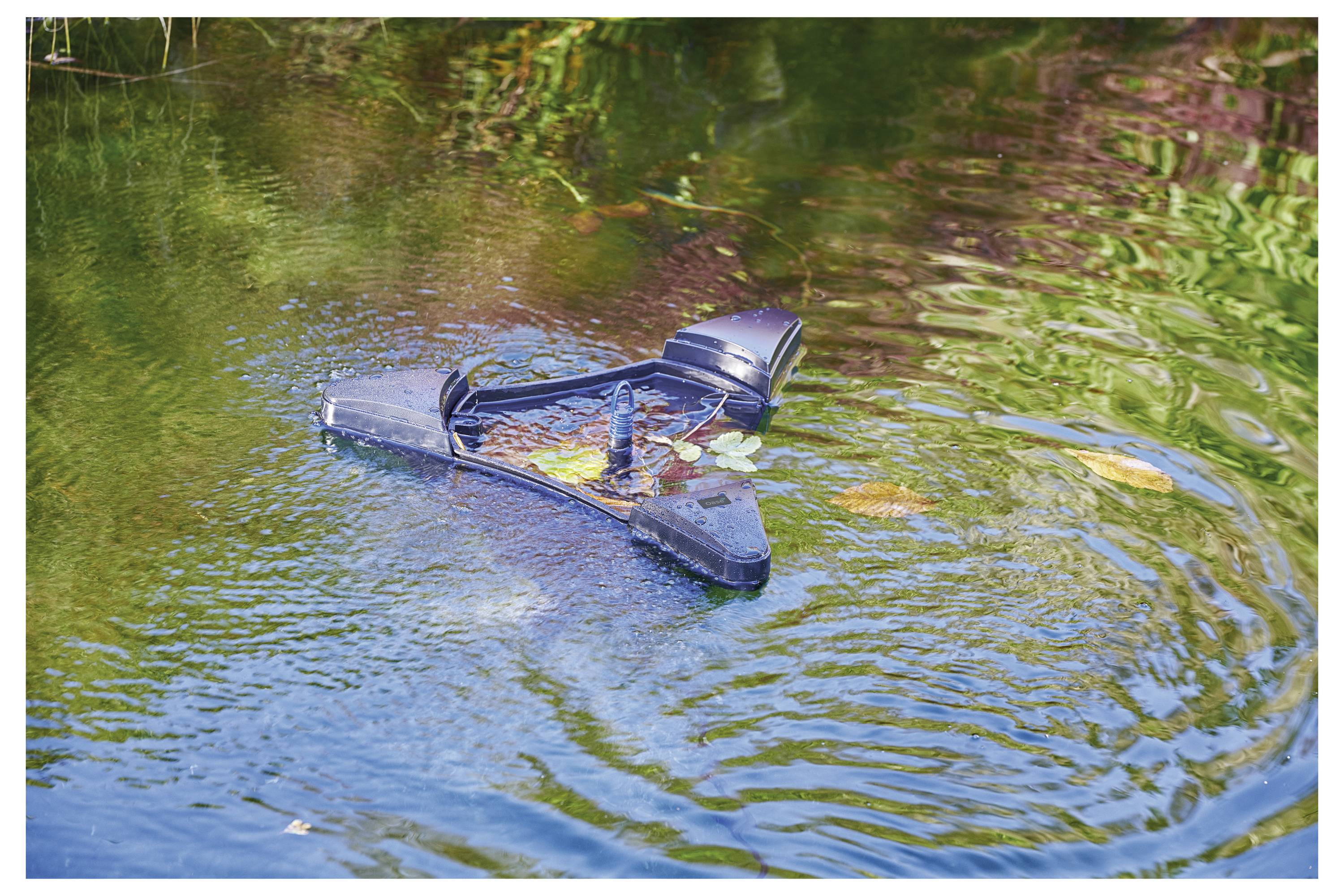 A broken side mirror floats on a green pond, partially submerged, surrounded by ripples and small patches of algae and plants.