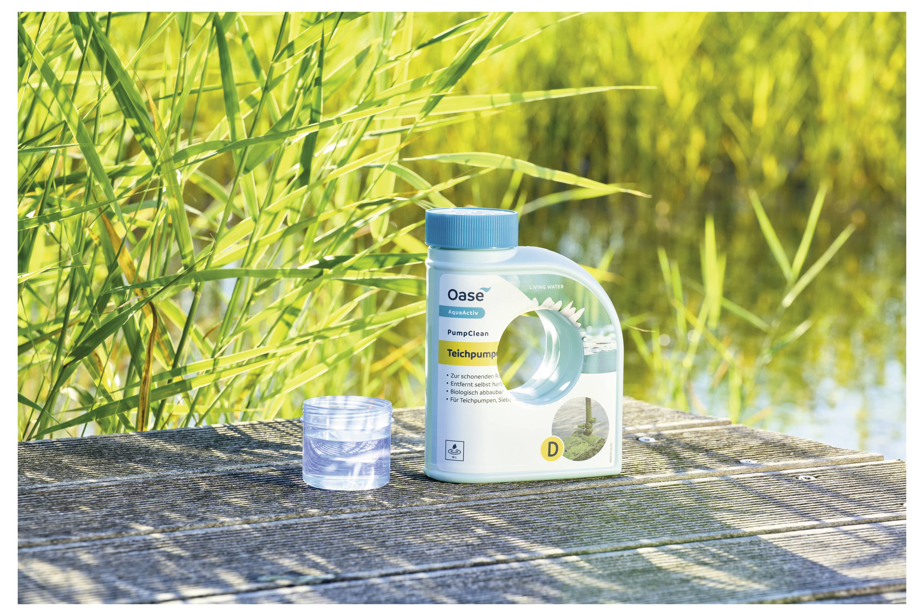 'Bottle of Oase pond cleaner and a clear plastic measuring cup on a wooden surface, with tall grass and water in the background.'