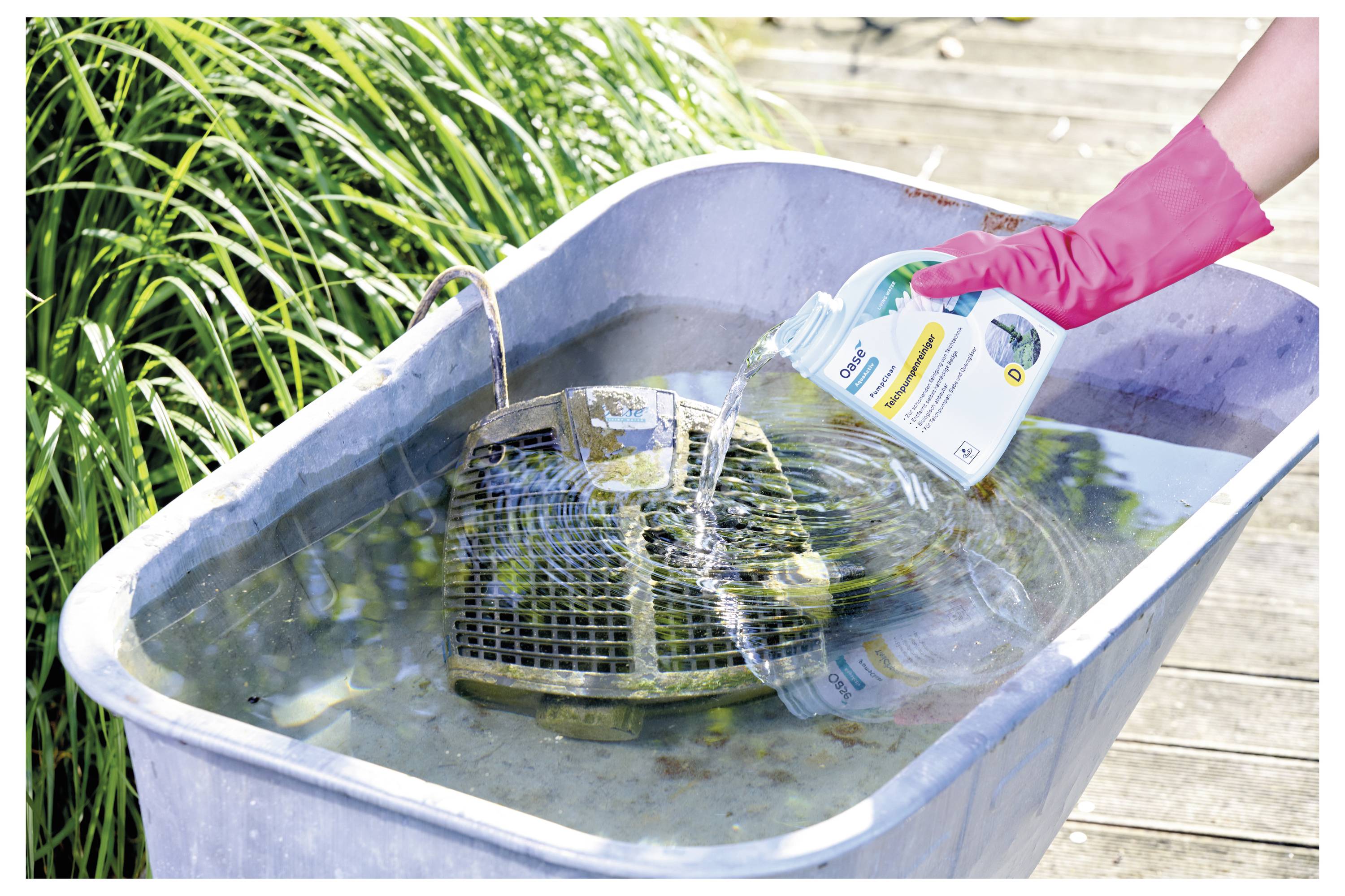 A person in a pink glove pours cleaner into a tub with a dirty grill grate soaking in water, next to grass and a wooden deck.