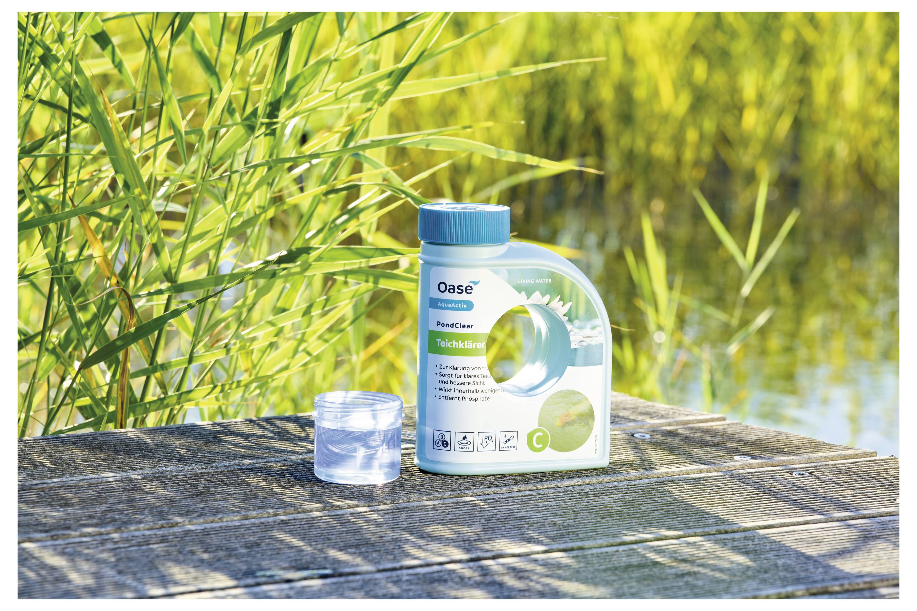 A container of 'Oase PondClear' on a wooden surface beside a clear measuring cup. Background features lush green reeds and water.