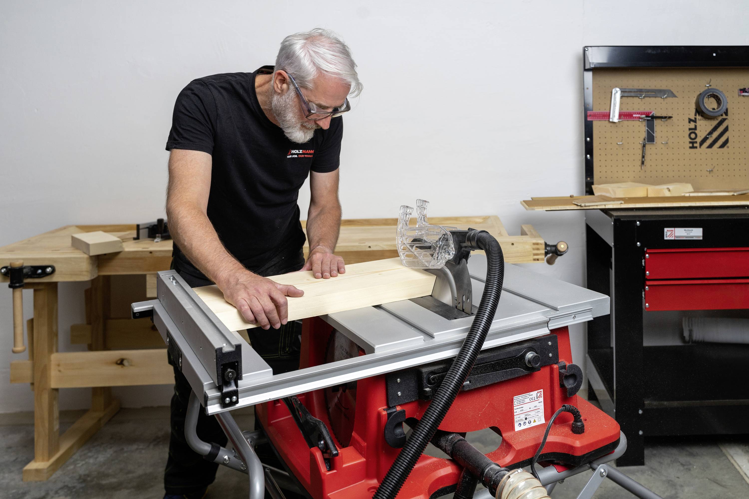 A person in a workshop is sawing a wooden board with a table saw. Tools can be seen mounted on a wall board in the background.