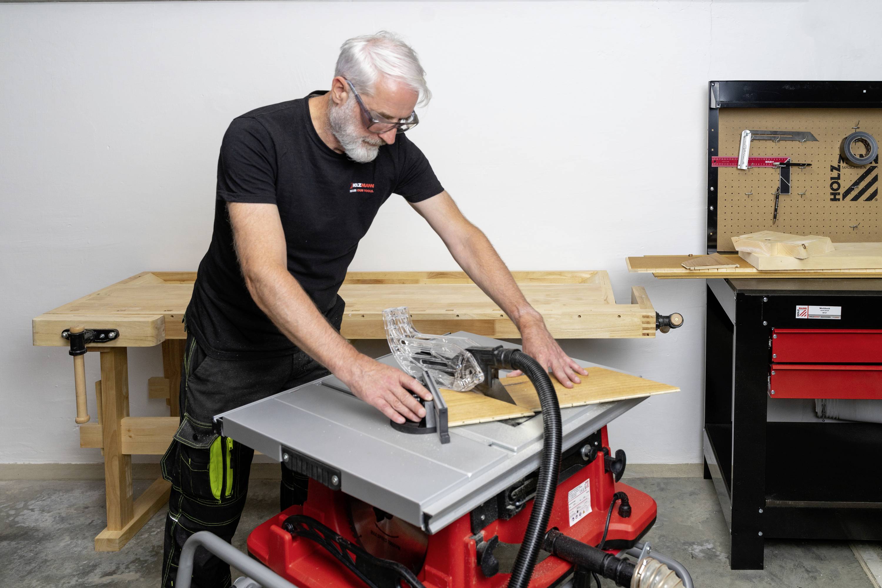 A person with white hair is working at a table saw in a workshop. Workbenches and tools can be seen in the background.