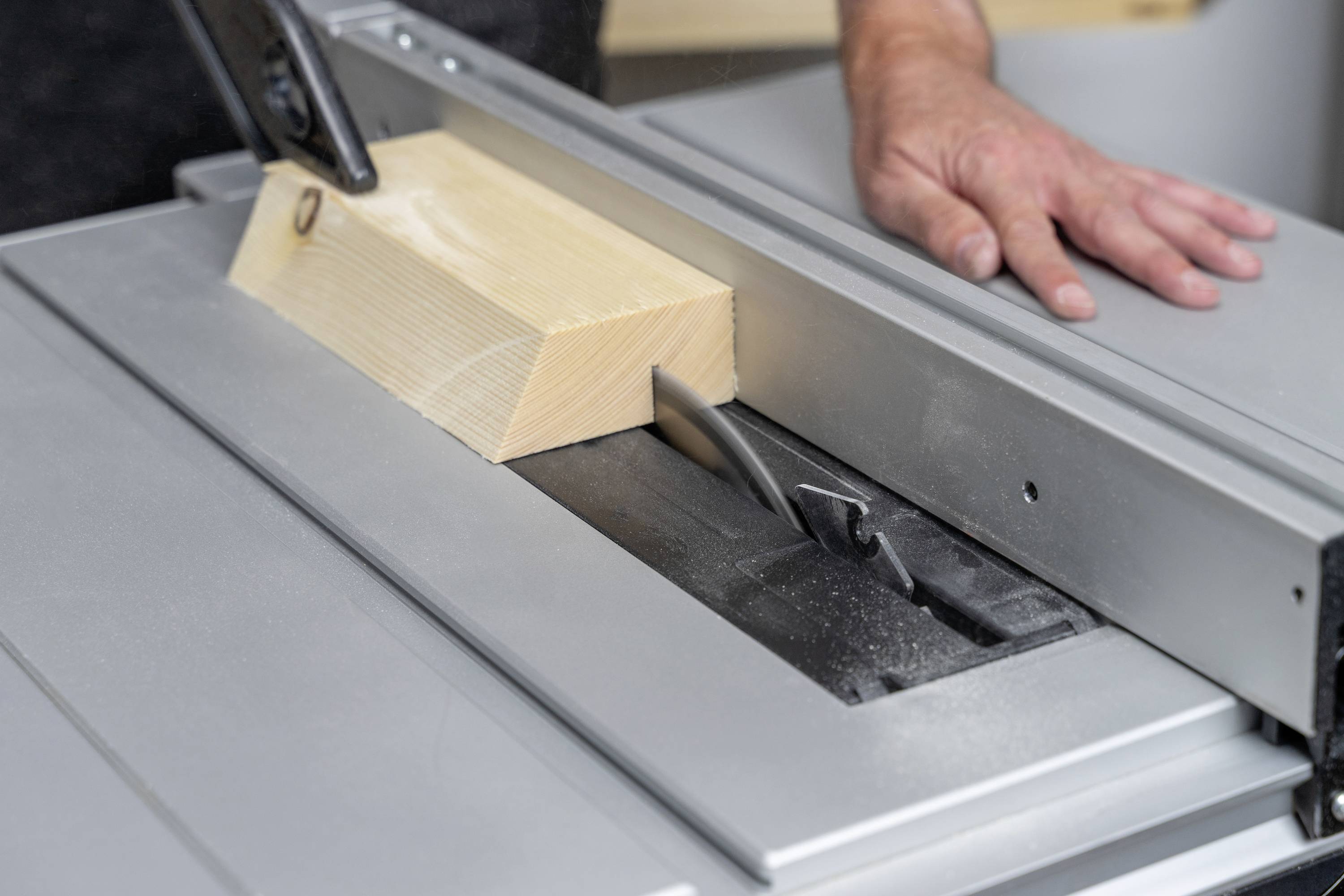 A hand carefully guides a wooden block over the saw blade of a table saw. Safety precautions must be observed.