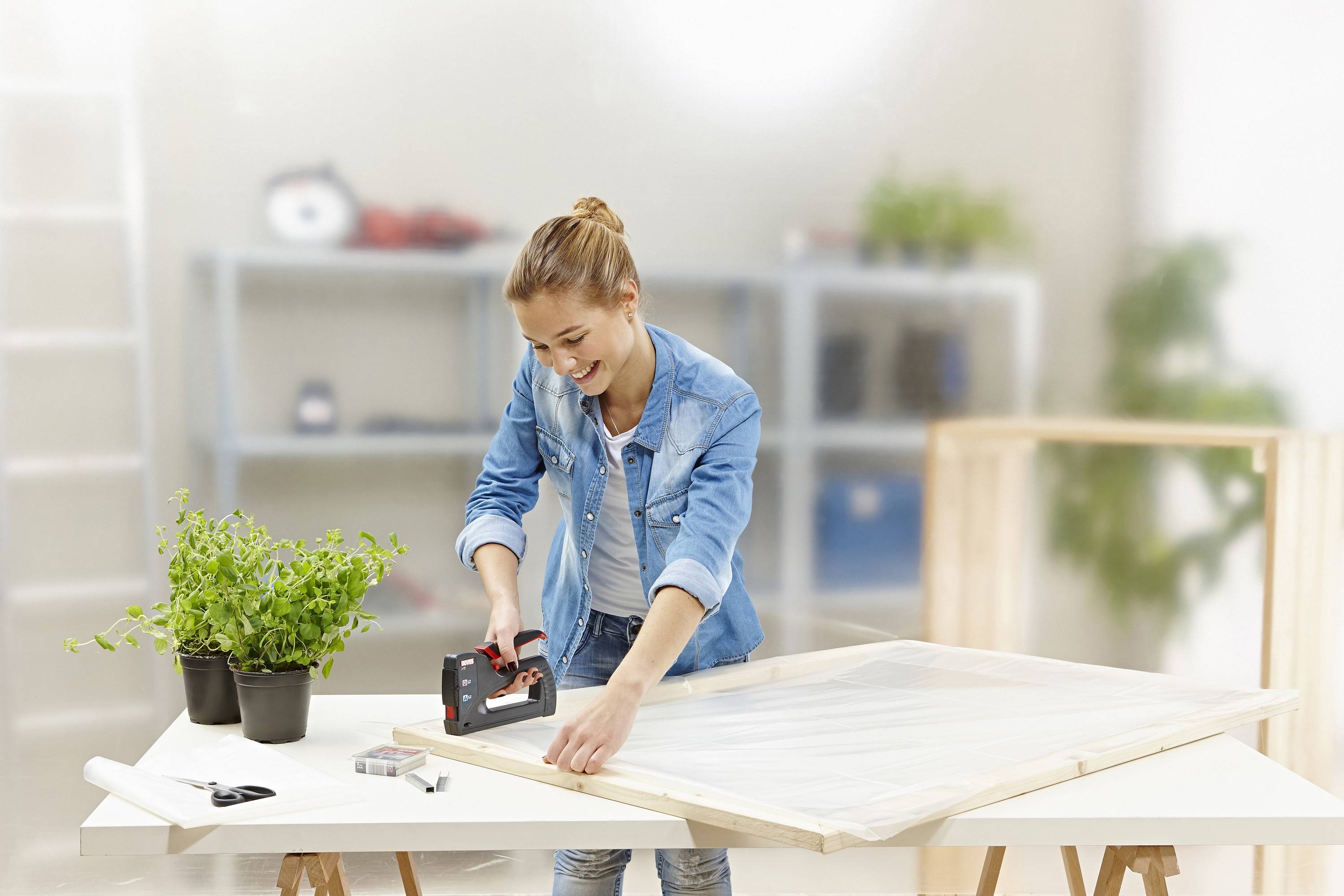 A woman is stapling fabric onto a wooden frame. Shelves, plants, and other work materials are visible in the background.