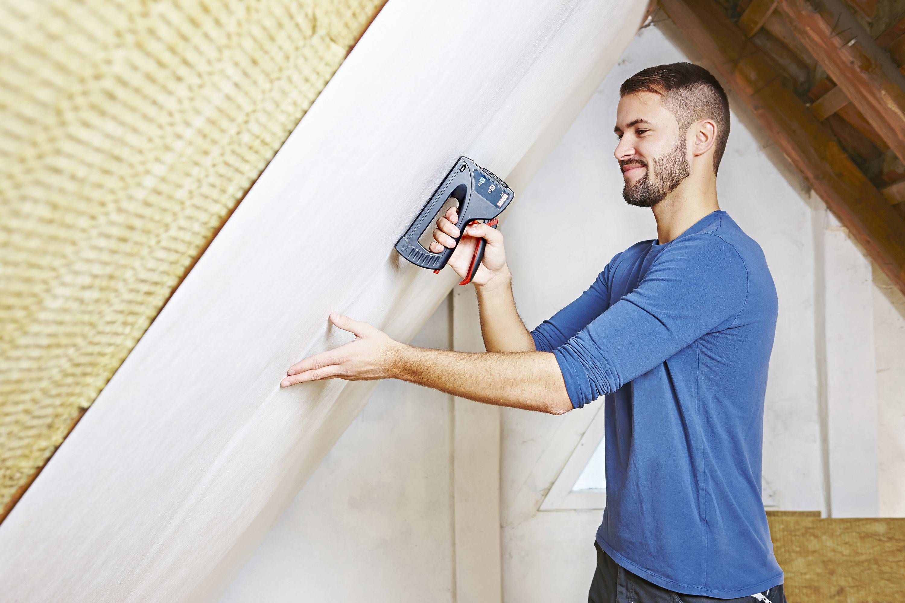 A man is firmly attaching an insulation board in an attic. He is wearing a blue shirt and working intently in the bright room.