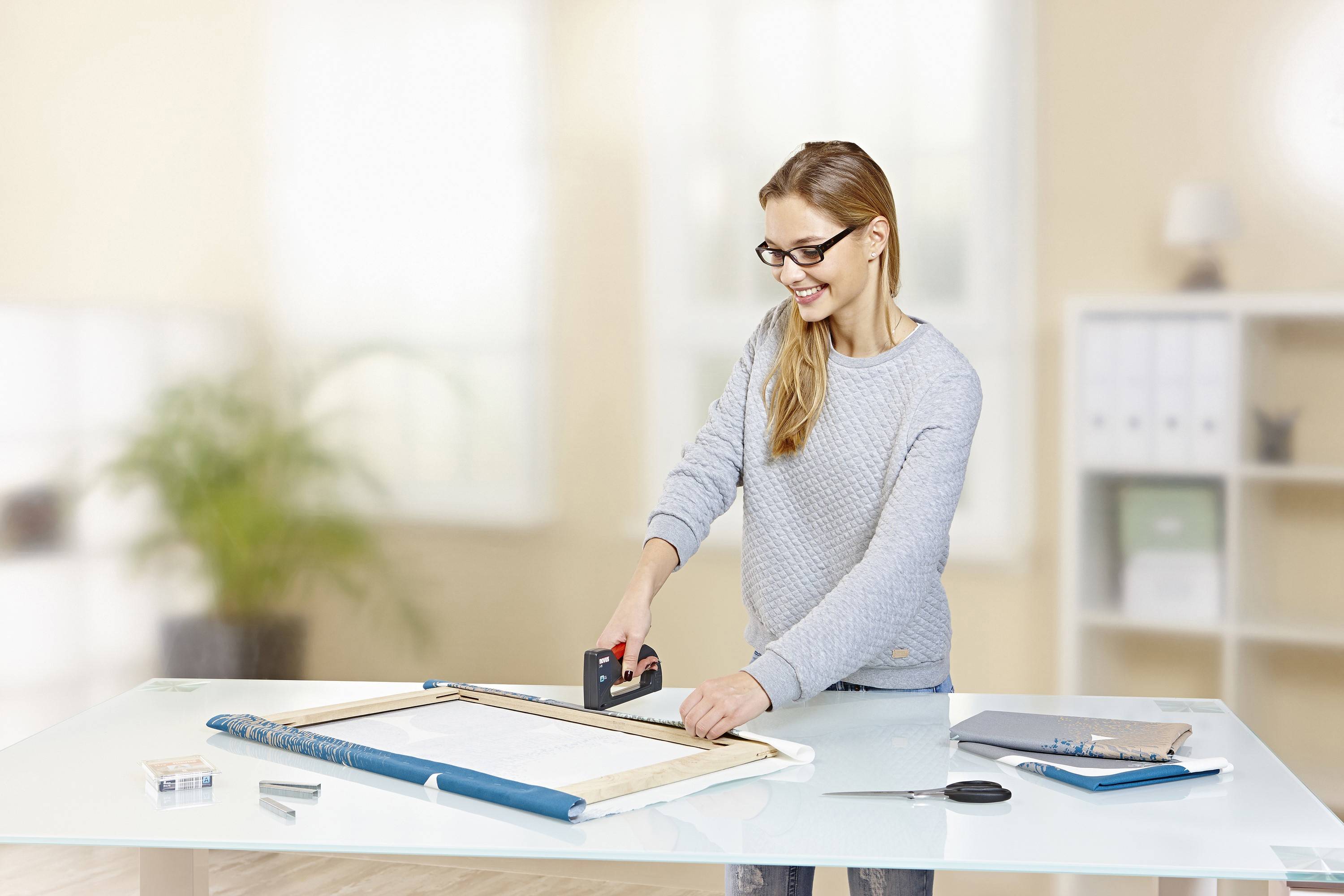 A woman is using a staple gun to attach a poster to a canvas. She is wearing glasses and smiling. Plants and folders can be seen in the background.