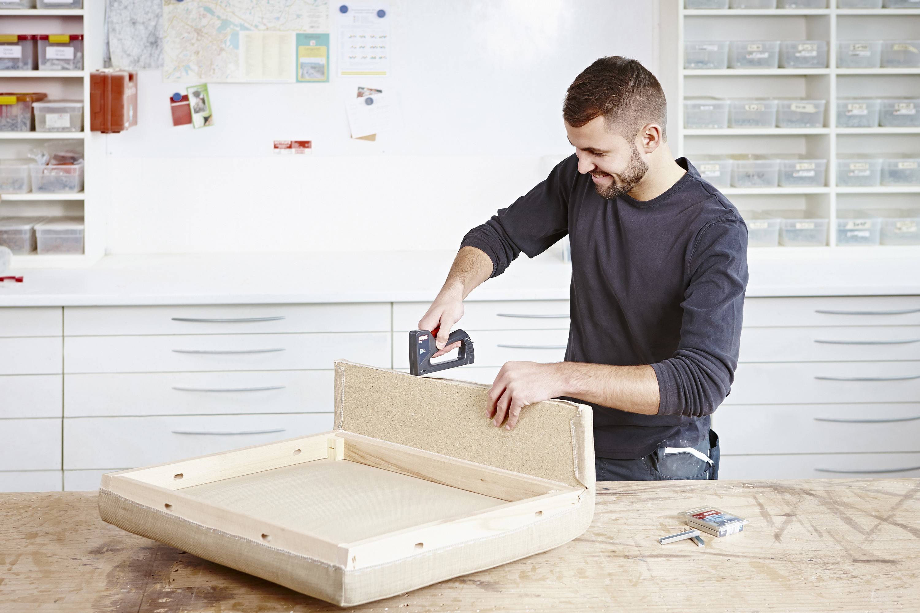 A man is working on a wooden floor covering in a workshop, using a staple gun to secure a fabric frame to a wooden floor.